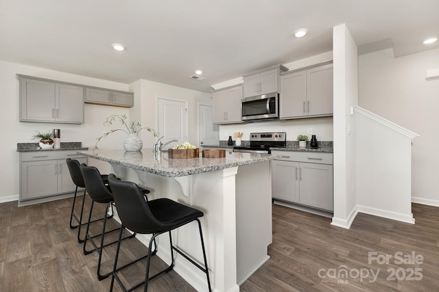 kitchen featuring appliances with stainless steel finishes, a center island with sink, gray cabinetry, a breakfast bar area, and light stone countertops