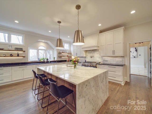 kitchen with a breakfast bar area, stainless steel stove, dark stone counters, an island with sink, and decorative light fixtures