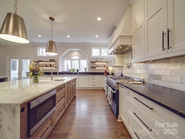 kitchen with white cabinets, appliances with stainless steel finishes, custom exhaust hood, dark stone counters, and decorative light fixtures