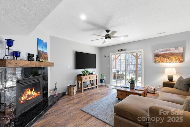 living area featuring visible vents, a tiled fireplace, ceiling fan, wood finished floors, and a textured ceiling