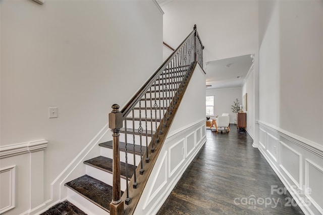 stairway featuring a towering ceiling, a wainscoted wall, wood finished floors, crown molding, and a decorative wall