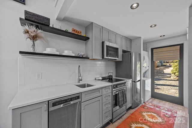 kitchen featuring stacked washer and clothes dryer, stainless steel appliances, gray cabinetry, open shelves, and a sink