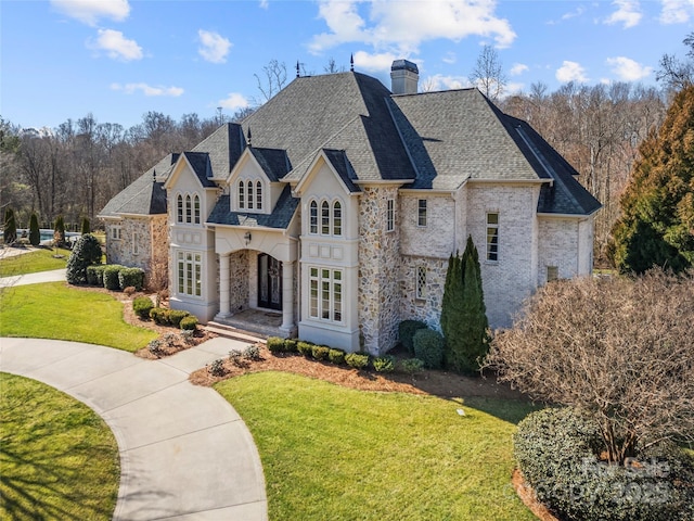 french country inspired facade featuring stone siding, roof with shingles, a chimney, and a front lawn