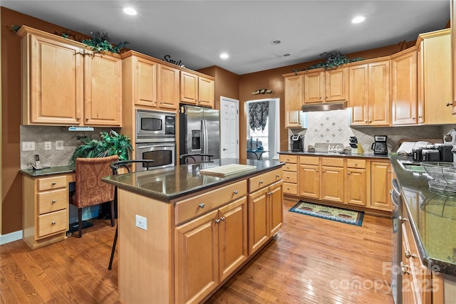 kitchen with under cabinet range hood, appliances with stainless steel finishes, backsplash, a center island, and light wood finished floors