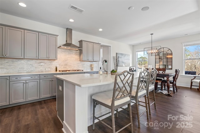 kitchen featuring visible vents, light countertops, wall chimney exhaust hood, an island with sink, and decorative light fixtures