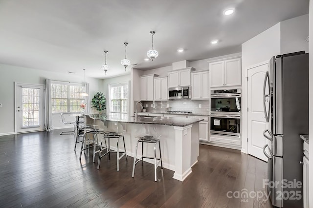 kitchen featuring decorative light fixtures, a center island with sink, appliances with stainless steel finishes, white cabinetry, and dark stone counters