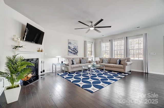 living room featuring ceiling fan, dark wood-type flooring, visible vents, baseboards, and a glass covered fireplace