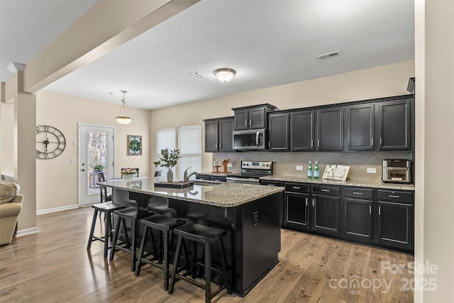 kitchen with light stone counters, a kitchen island with sink, stainless steel appliances, a breakfast bar, and hanging light fixtures