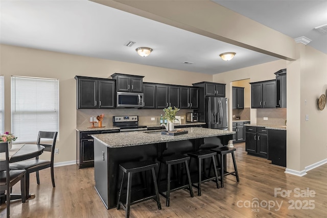 kitchen with stainless steel appliances, light stone counters, a center island with sink, and dark cabinets