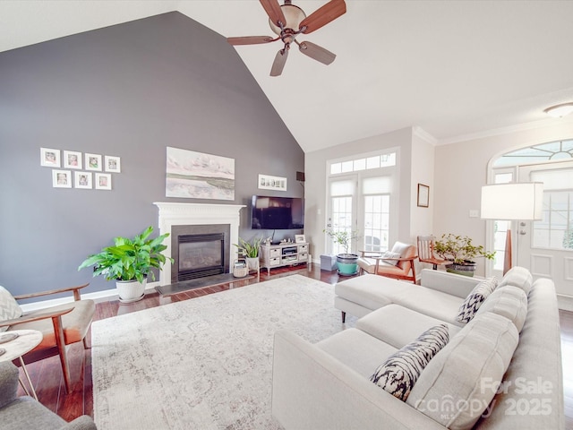 living area with a ceiling fan, a glass covered fireplace, wood finished floors, high vaulted ceiling, and baseboards