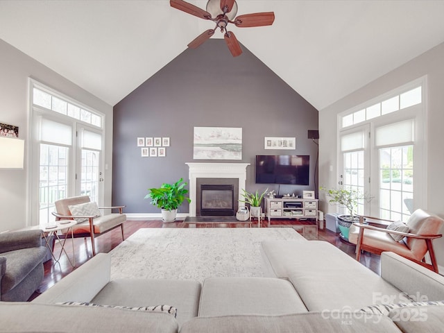 living room with baseboards, a glass covered fireplace, ceiling fan, wood finished floors, and high vaulted ceiling