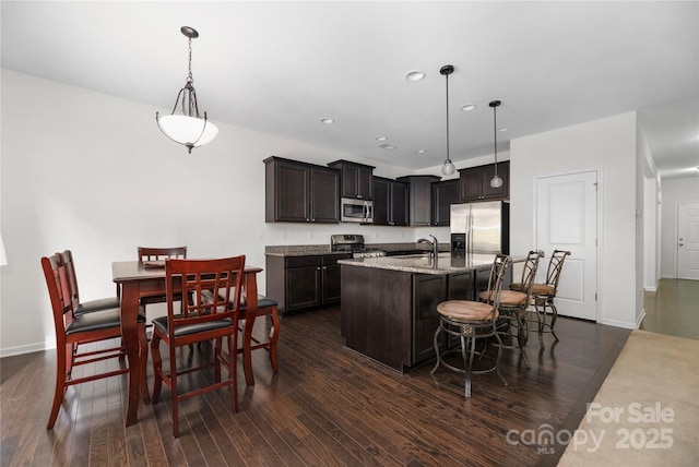 kitchen featuring a kitchen island with sink, appliances with stainless steel finishes, hanging light fixtures, dark brown cabinetry, and dark wood-style flooring