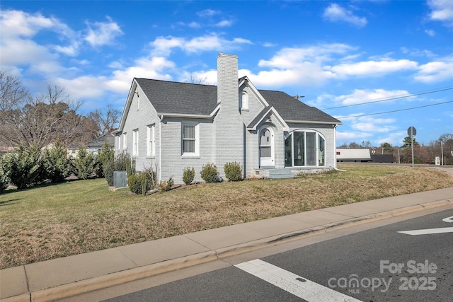 view of front facade featuring brick siding, central air condition unit, a front yard, roof with shingles, and a chimney