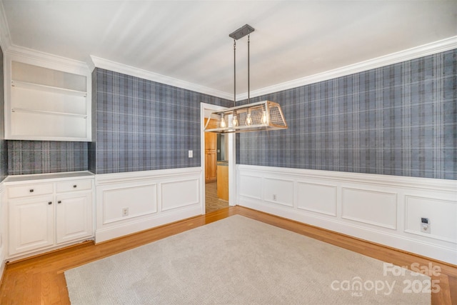 unfurnished dining area with a wainscoted wall, light wood-style floors, ornamental molding, and wallpapered walls