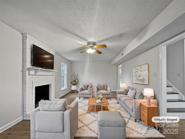 living area featuring baseboards, dark wood finished floors, ceiling fan, stairs, and a brick fireplace