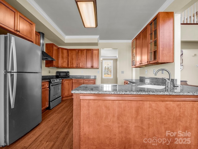 kitchen with dark countertops, appliances with stainless steel finishes, glass insert cabinets, wall chimney range hood, and a peninsula