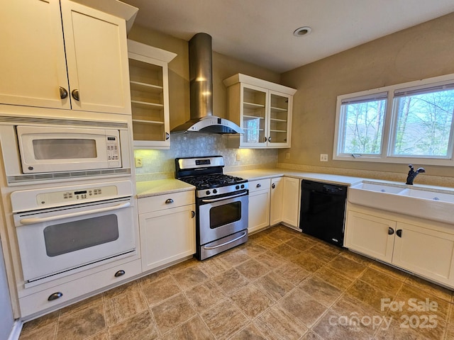 kitchen featuring white appliances, glass insert cabinets, light countertops, wall chimney range hood, and a sink