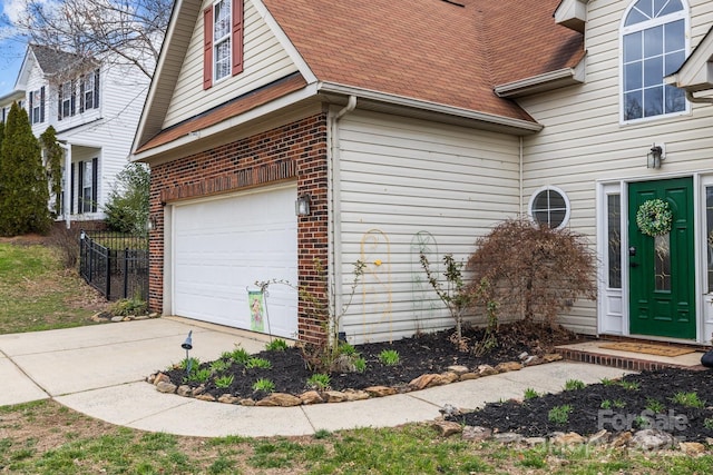 view of exterior entry featuring brick siding, a shingled roof, fence, driveway, and an attached garage