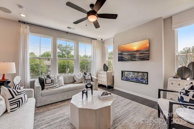 living area featuring dark wood-style floors, visible vents, a ceiling fan, a glass covered fireplace, and baseboards