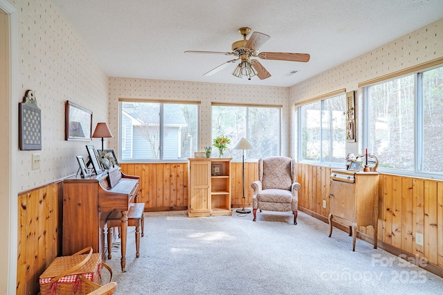 sitting room featuring wallpapered walls, wainscoting, ceiling fan, carpet, and a textured ceiling