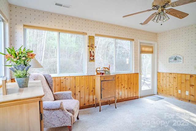 living area featuring ceiling fan, a wainscoted wall, carpet flooring, visible vents, and wallpapered walls