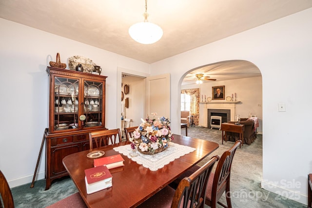 dining area featuring arched walkways, ceiling fan, a fireplace, baseboards, and dark carpet