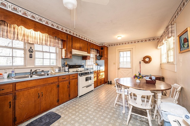 kitchen with white gas stove, light countertops, brown cabinetry, a sink, and under cabinet range hood