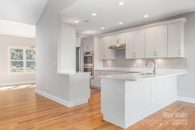 kitchen with light countertops, white cabinetry, a sink, a peninsula, and under cabinet range hood