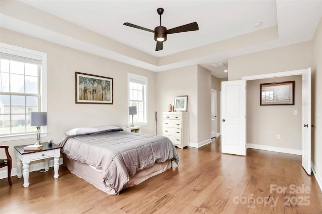 bedroom featuring a tray ceiling, multiple windows, baseboards, and light wood-type flooring