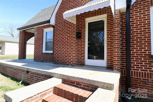 view of exterior entry with a shingled roof and brick siding