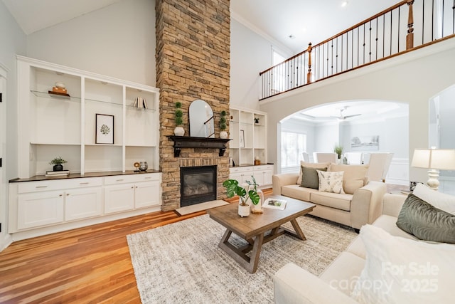 living area featuring arched walkways, ornamental molding, a stone fireplace, a towering ceiling, and light wood-type flooring