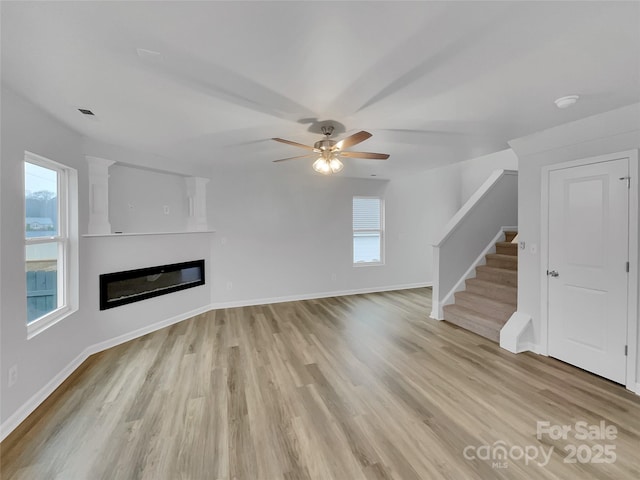 unfurnished living room featuring light wood-style floors, a glass covered fireplace, a ceiling fan, baseboards, and stairs