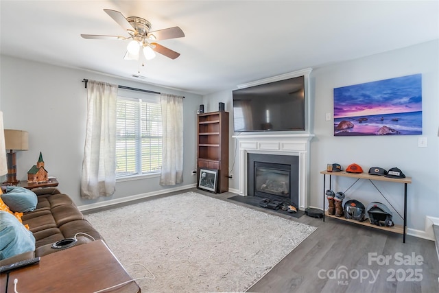 living room featuring a fireplace with flush hearth, a ceiling fan, baseboards, and wood finished floors