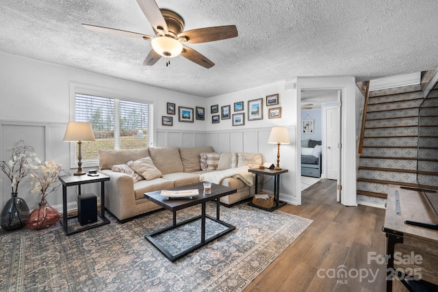 living area with a wainscoted wall, ceiling fan, stairway, wood finished floors, and a textured ceiling