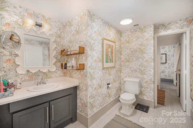 bathroom with toilet, wallpapered walls, baseboards, and vanity