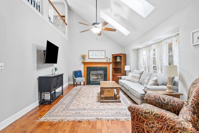 living area with a skylight, baseboards, a glass covered fireplace, ceiling fan, and wood finished floors