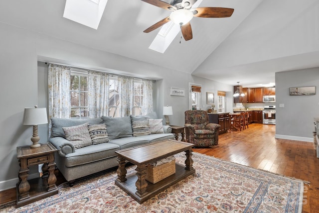 living room with high vaulted ceiling, a skylight, a ceiling fan, baseboards, and light wood finished floors