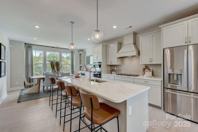 kitchen featuring pendant lighting, a center island with sink, stainless steel appliances, light stone countertops, and premium range hood