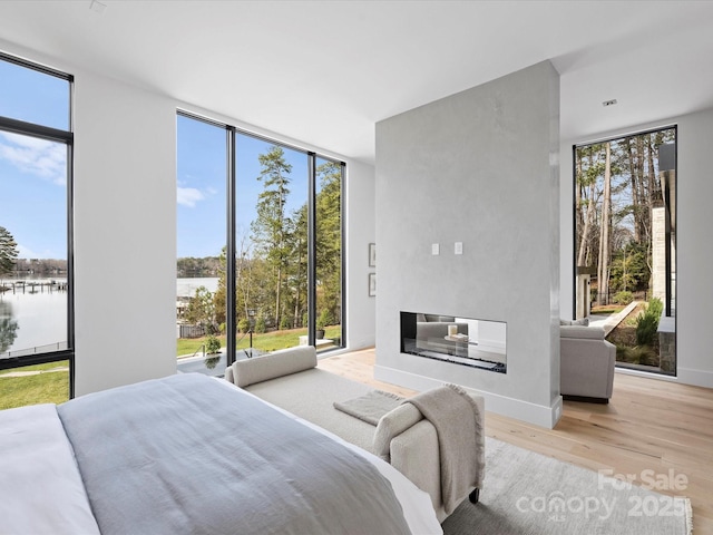 bedroom featuring baseboards, light wood-style flooring, a water view, a wall of windows, and a multi sided fireplace