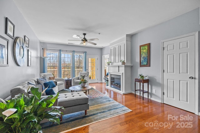 living area featuring a glass covered fireplace, ceiling fan, baseboards, and wood finished floors