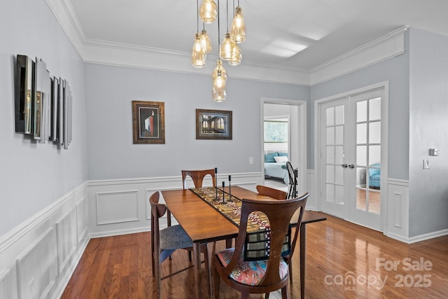 dining area with a decorative wall, a wainscoted wall, wood finished floors, french doors, and crown molding