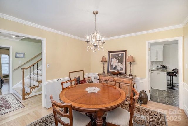 dining room with a wainscoted wall, stairway, light wood-style flooring, an inviting chandelier, and ornamental molding