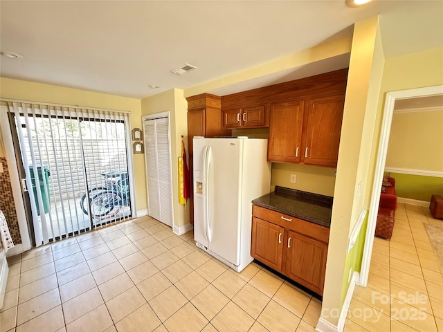 kitchen with light tile patterned floors, white refrigerator with ice dispenser, visible vents, brown cabinetry, and dark stone counters