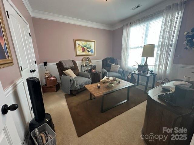 living area featuring visible vents, a decorative wall, ornamental molding, wainscoting, and carpet flooring