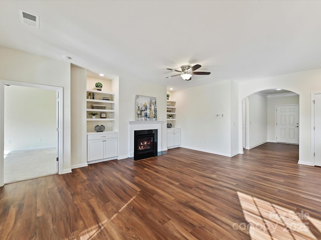 unfurnished living room with a warm lit fireplace, built in shelves, dark wood-type flooring, and baseboards