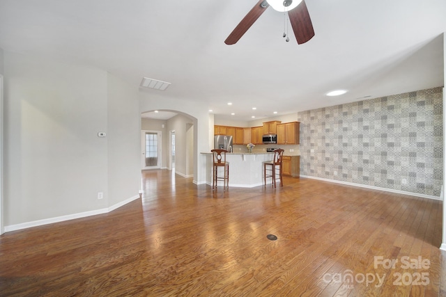 unfurnished living room with wood finished floors, visible vents, baseboards, wallpapered walls, and arched walkways