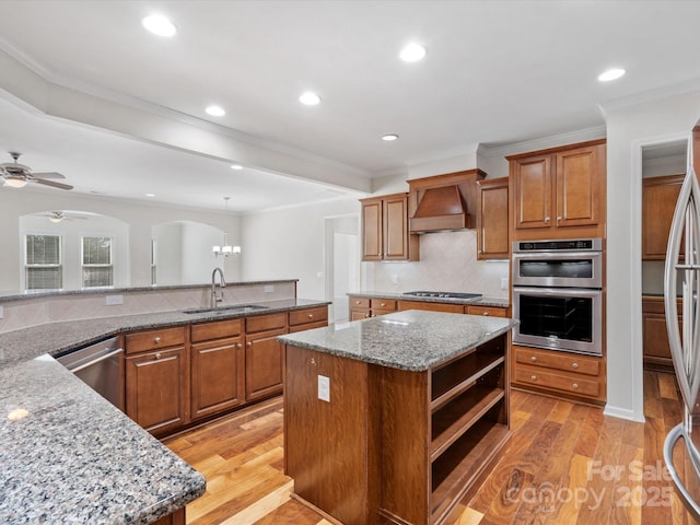 kitchen featuring brown cabinetry, a kitchen island, stainless steel appliances, premium range hood, and a sink