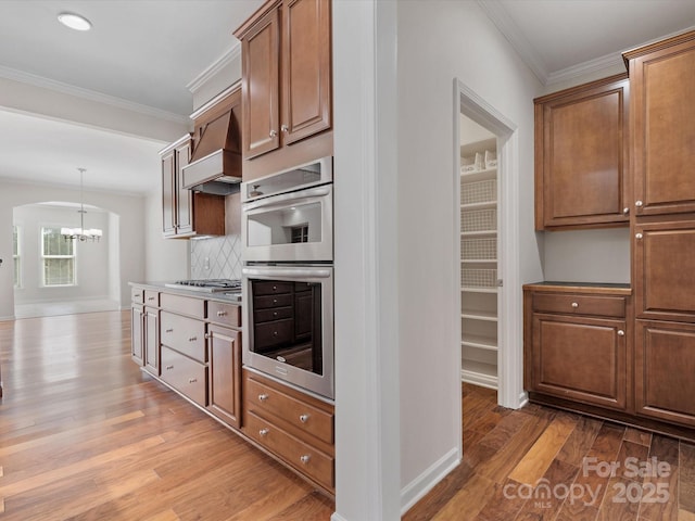 kitchen featuring stainless steel appliances, custom range hood, backsplash, light wood finished floors, and crown molding