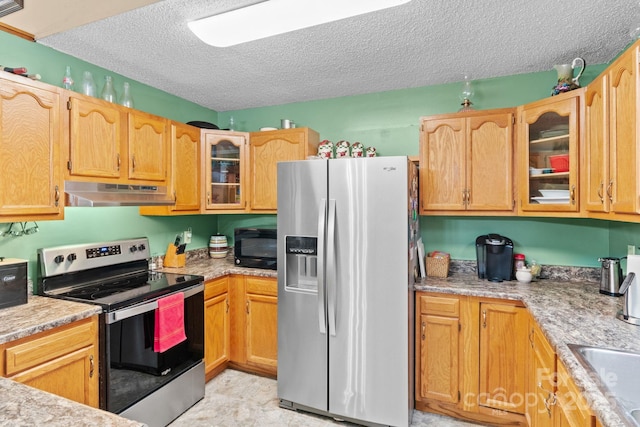 kitchen with light stone counters, appliances with stainless steel finishes, glass insert cabinets, a textured ceiling, and under cabinet range hood