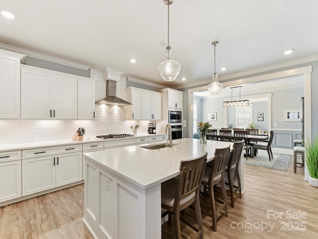 kitchen featuring a center island with sink, stainless steel appliances, light countertops, wall chimney range hood, and a sink